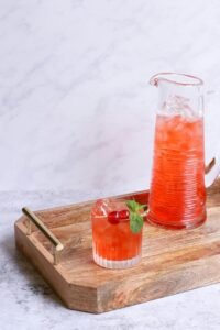 Cherry and mint flavored beverage served in glass and jug on wooden tray.
